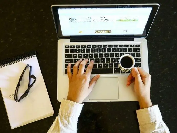 Foto de uma pessoa trabalhando no laptop e segurando uma xícara de café. Do lado esquerdo há um bloco de notas com um óculos em cima. A foto está cortada e não é possível ver o rosto da pessoa